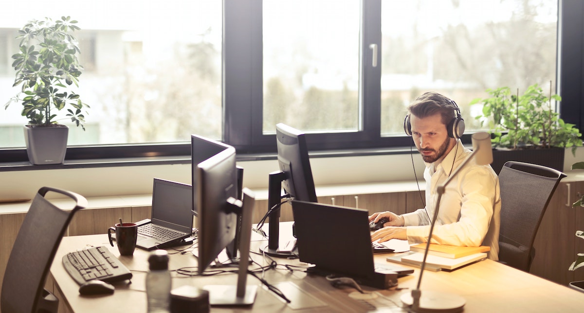 Man wearing headphones and working at a desk with two monitors in an office 