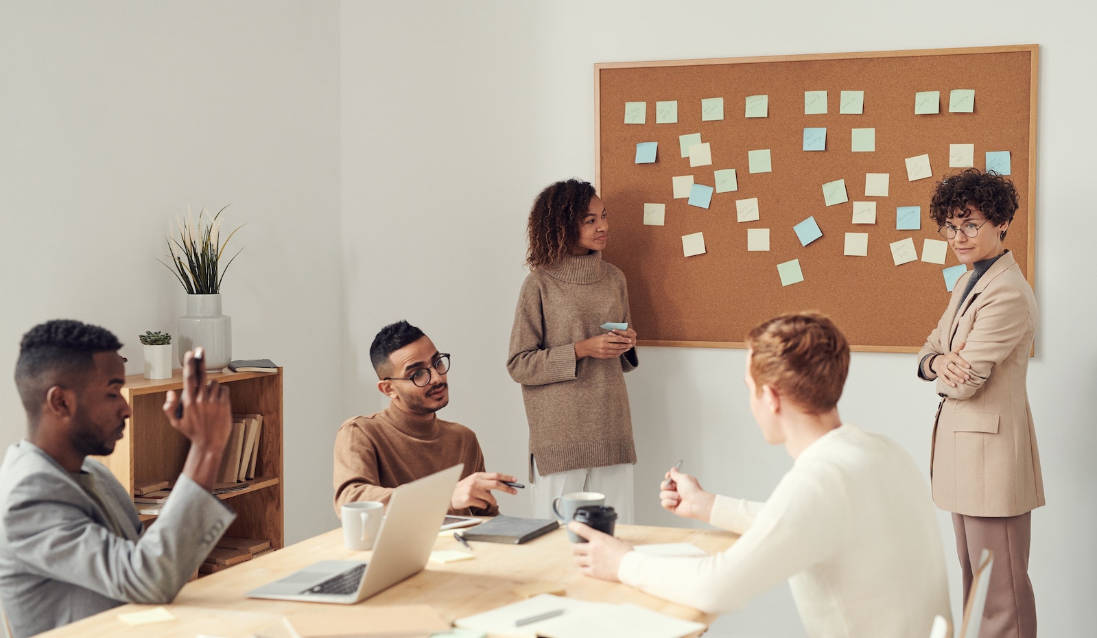 Five colleagues in a meeting brainstorming and planning with sticky notes on a cork board. 