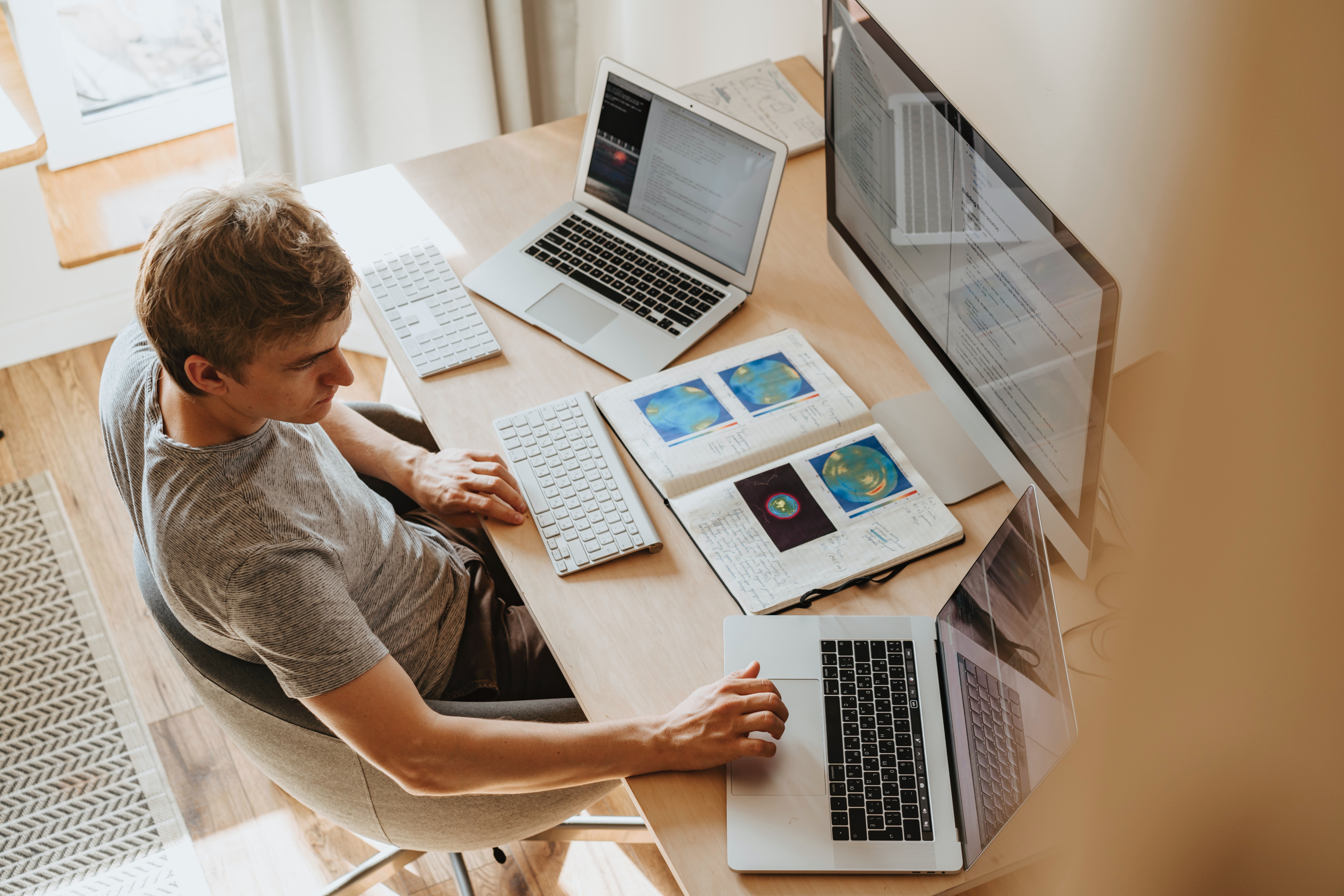 Person with short hair working at a desk with two laptops, a monitor, and a notebook. 